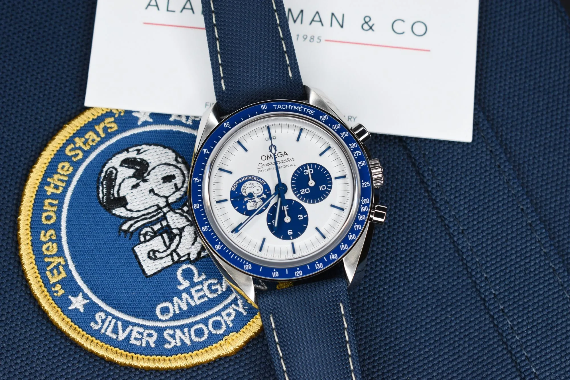 Omega Speedmaster Silver Snoopy Award with a silver dial, blue subdials and a blue ceramic bezel on a blue fabric strap, resting on a Silver Snoopy patch and an Alan Furman & Co card